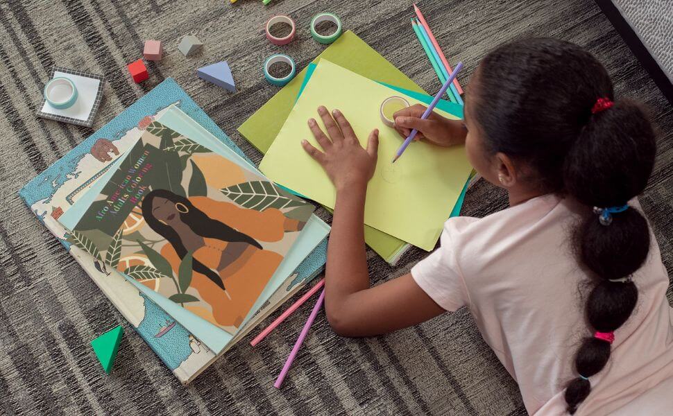 A girl coloring on yellow paper next to an Afro-American women coloring book, surrounded by craft supplies.