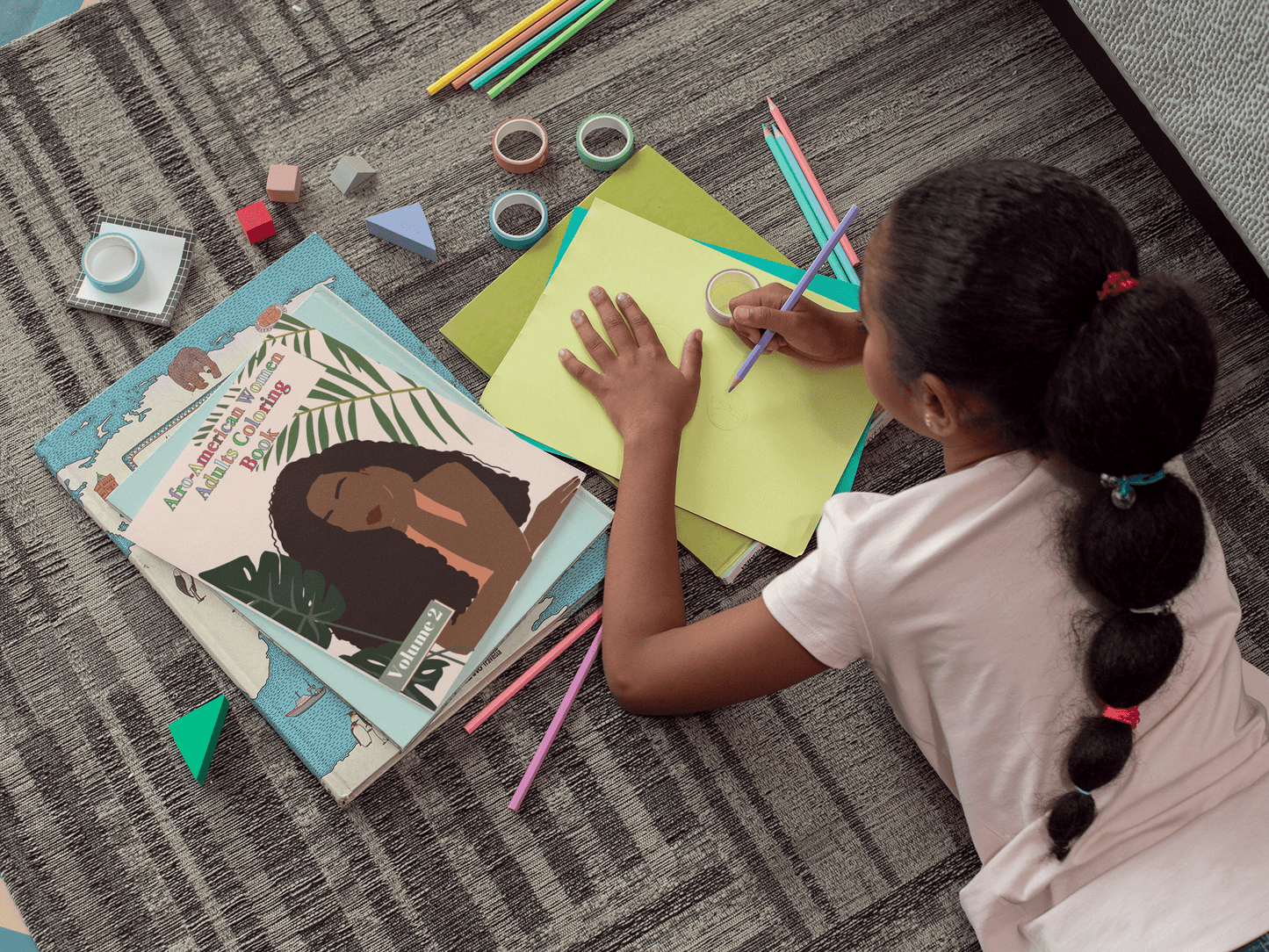 Child coloring on a green sheet with Afro-American Women Adults Coloring Book Volume 2 beside colorful art supplies.