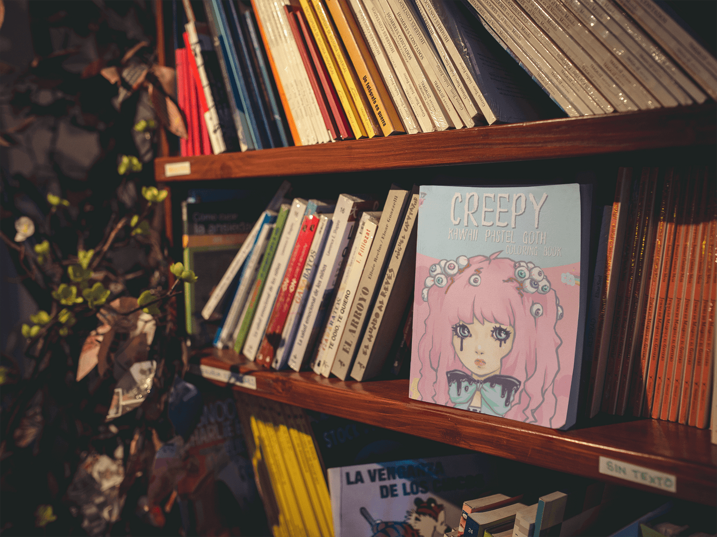 Creepy Kawaii Pastel Goth Coloring Book displayed on a bookshelf among various colorful books and plant decor.