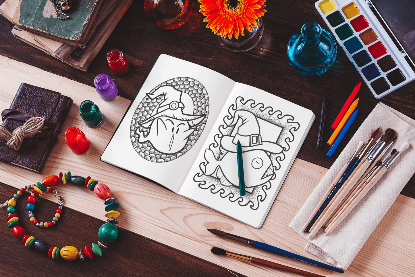 Open coloring book featuring spooky kawaii designs, surrounded by art supplies and colorful paints on a wooden table.