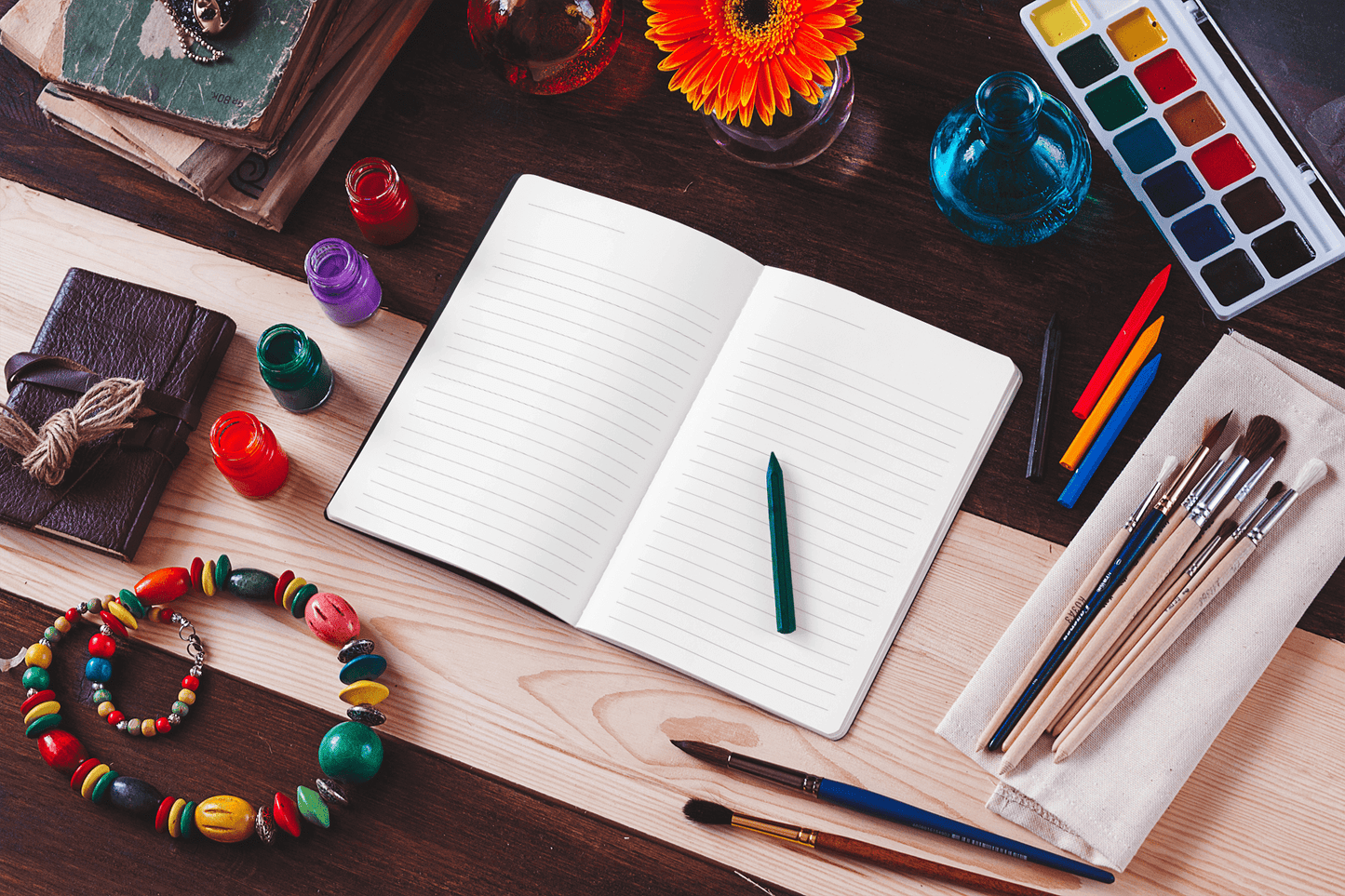 Open blank pages of a journal among art supplies, beads, paints, and flowers on a wooden table.