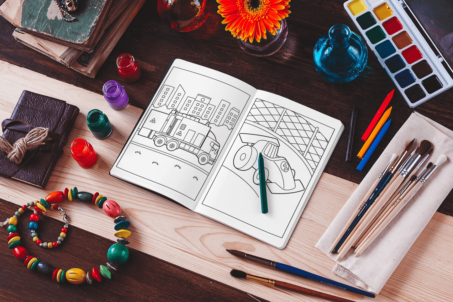 Open coloring book showing vehicle illustrations with art supplies on a wooden table.