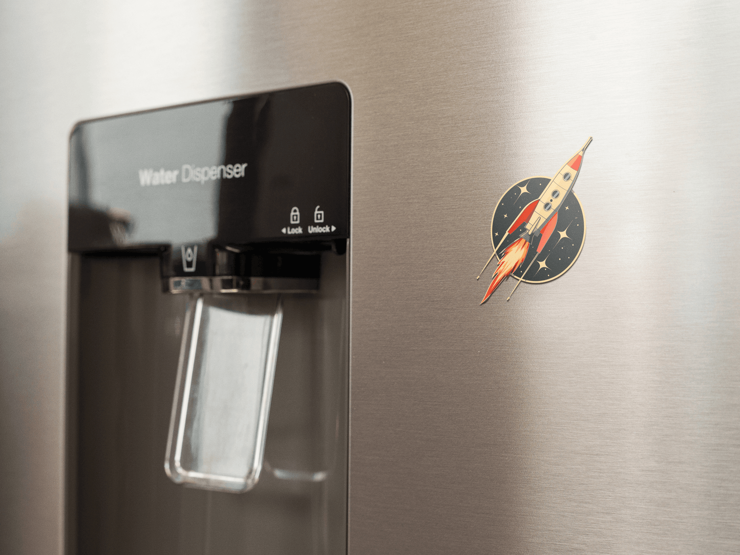 Atomic Age spacecraft sticker on a stainless steel refrigerator next to water dispenser.