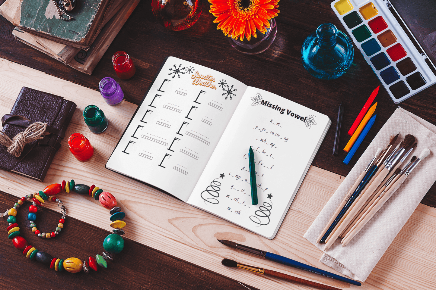 Open game book page with 'Missing Vowel' puzzle, surrounded by art supplies and colorful objects on a wooden table.