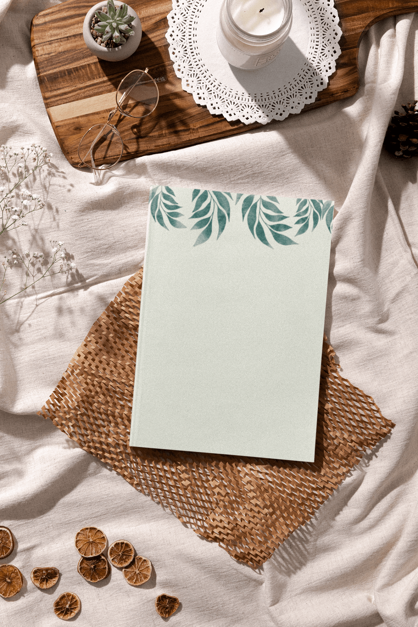 A minimalist notebook with a leafy design on a woven mat, surrounded by dried citrus and decorative items.