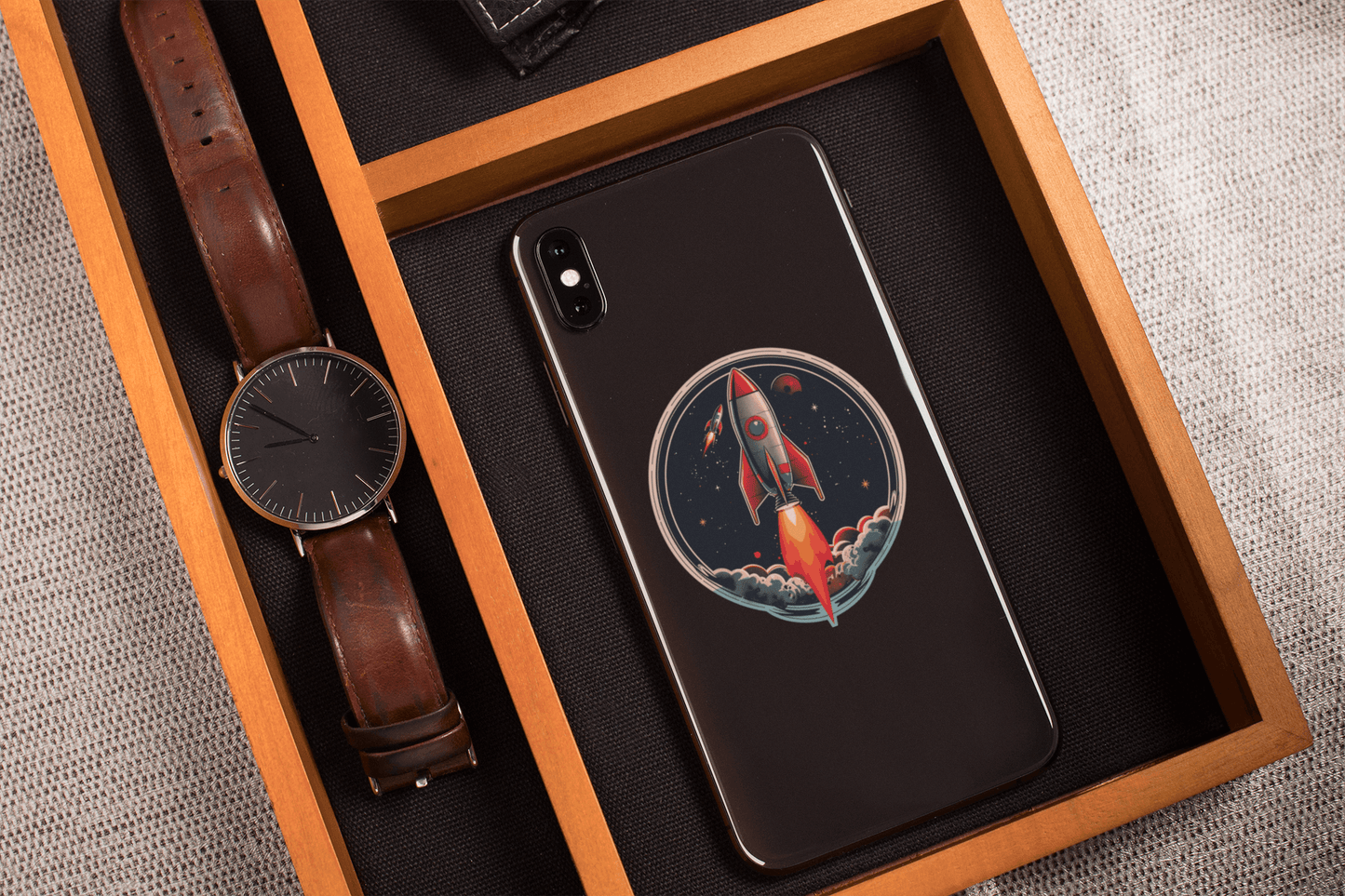 Atomic Age spacecraft sticker on a smartphone resting on a wooden tray with a watch.