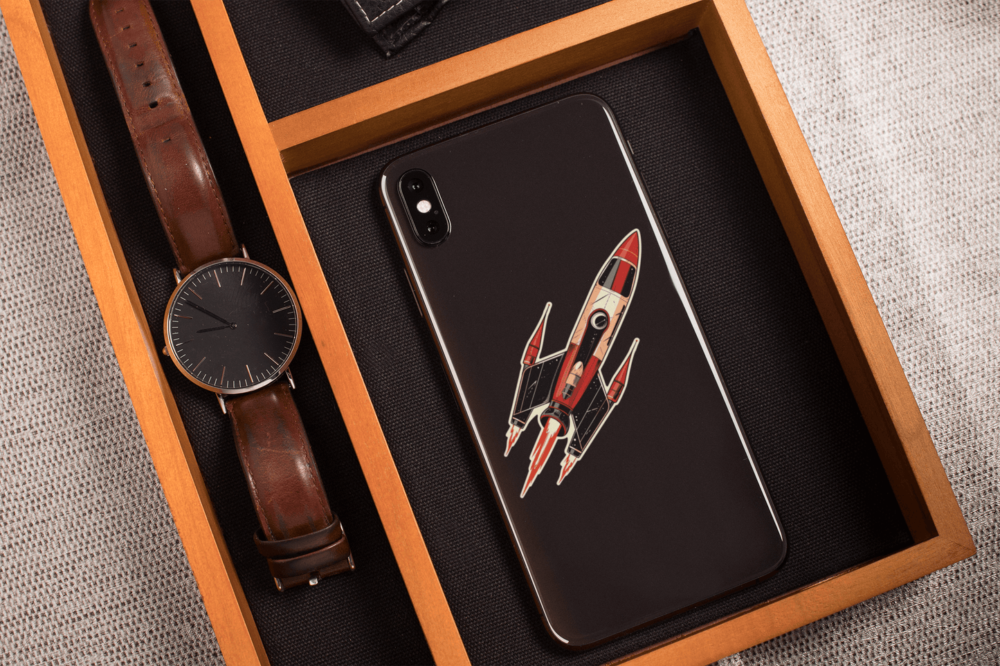 Atomic Age spacecraft sticker on a smartphone in a wooden tray with a watch.