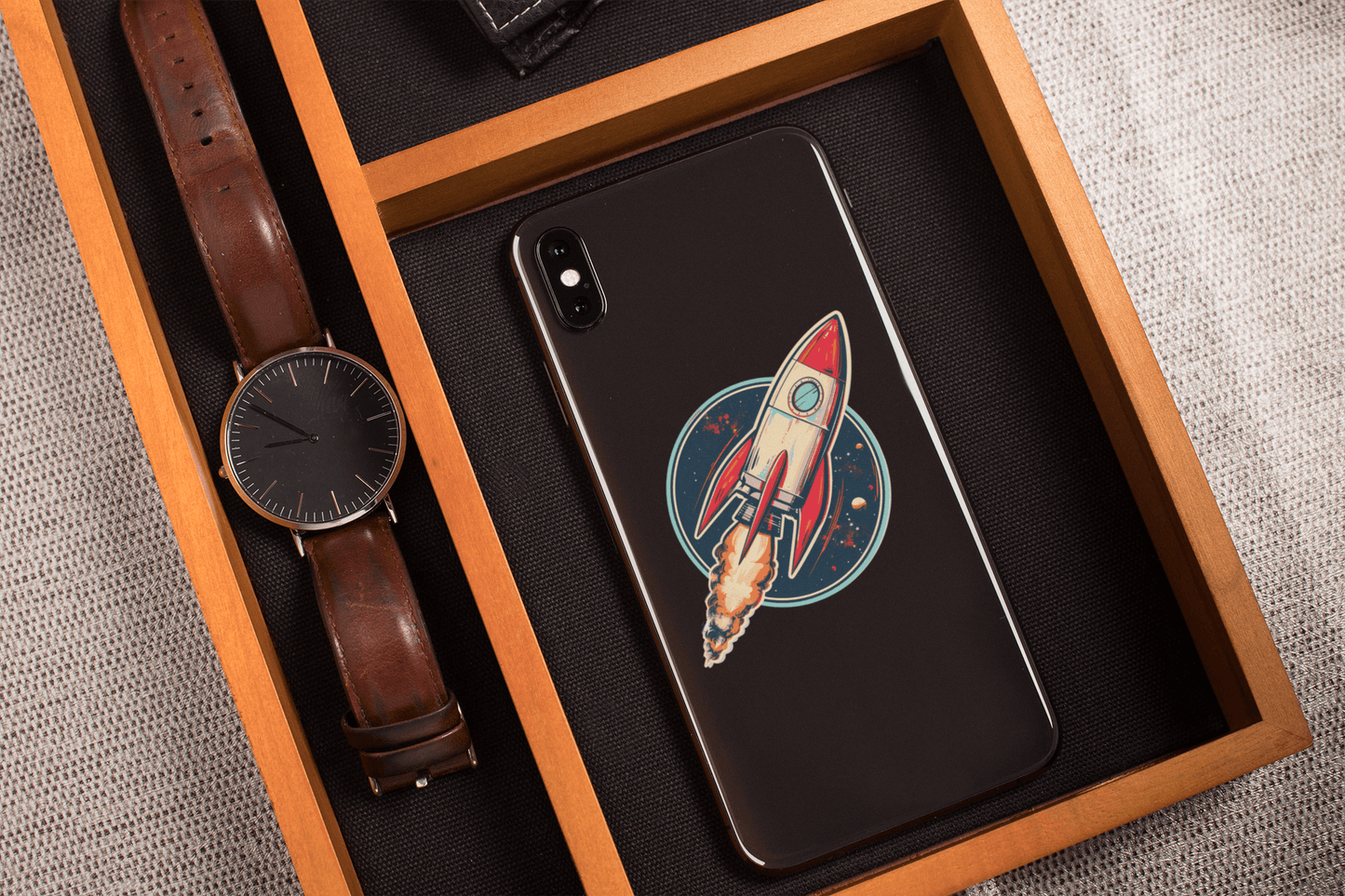 Atomic Age spacecraft sticker on a phone case beside a watch and a textured fabric background.