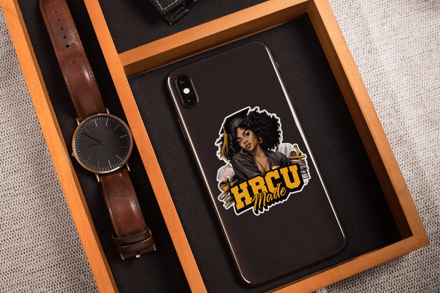 HBCU Made sticker on a smartphone case showcased in a wooden tray with a watch and accessories.