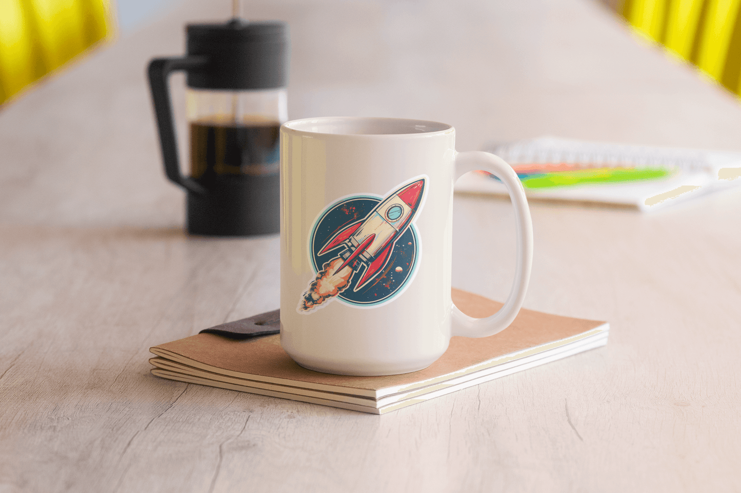 Atomic Age spacecraft sticker on a white mug placed on a wooden table next to a notebook.