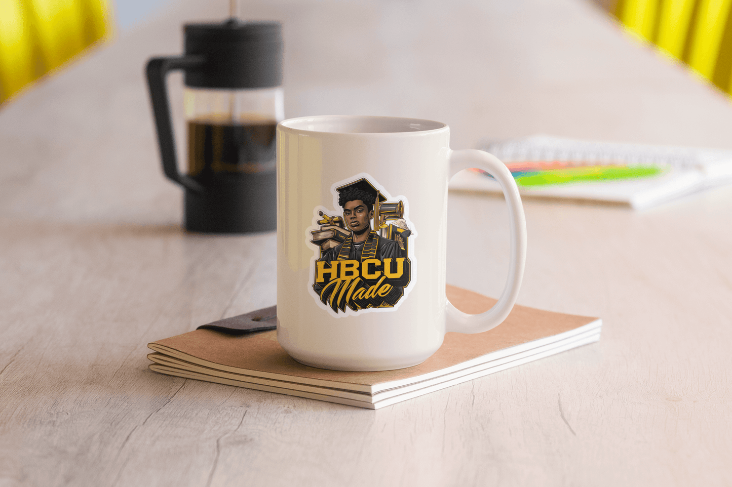 HBCU Made sticker on a white mug placed on a wooden table with a notebook and coffee maker in the background.