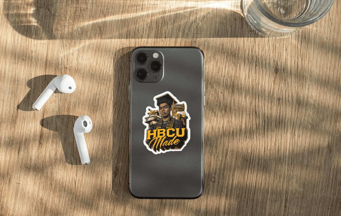 HBCU Made sticker on a smartphone with white earbuds and a glass of water in natural light.
