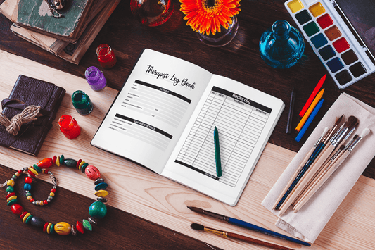 Therapist log book open on a wooden table with colorful paints, brushes, and floral decor surrounding it.