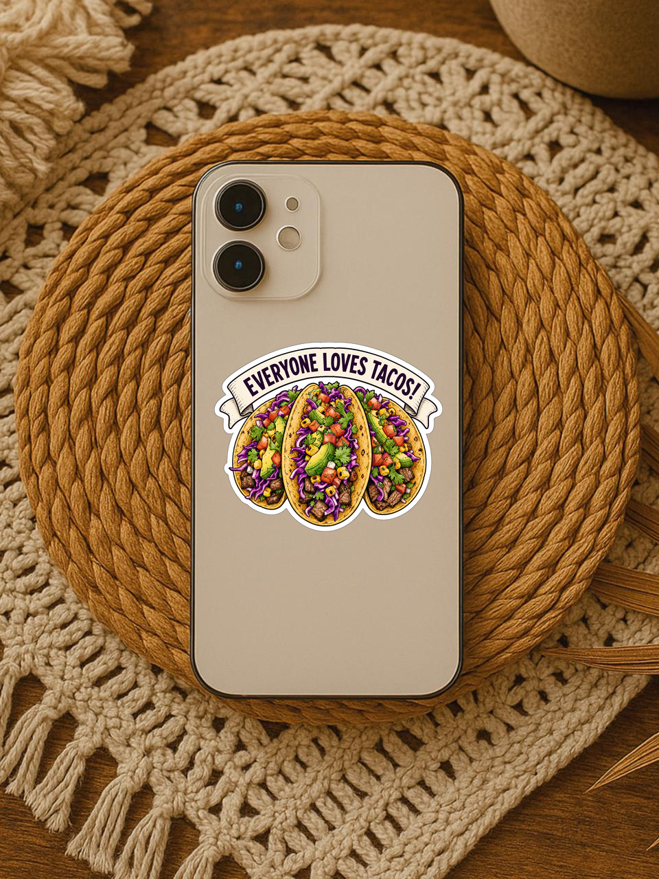 Mockup showing the Everyone Loves Tacos! sticker design on the back of a beige phone resting on a woven placemat.