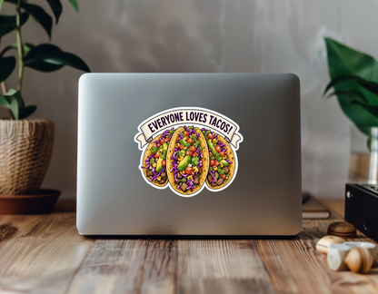 Mockup showing the Everyone Loves Tacos! sticker design on the back of a laptop between houseplants on a wooden desk.
