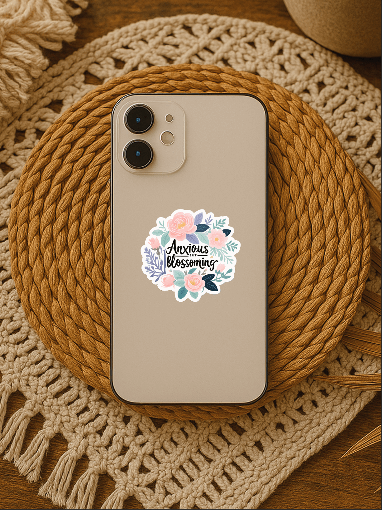 Anxious But Blossoming pastel floral affirmation vinyl sticker on a phone resting on a decorative woven mat.