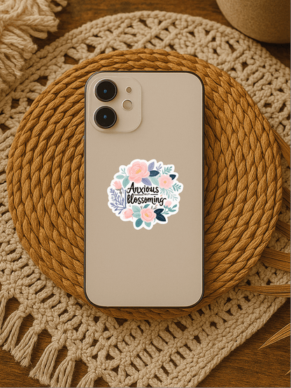 Anxious But Blossoming pastel floral affirmation vinyl sticker on a phone resting on a decorative woven mat.